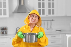 Young woman in raincoat collecting leaking water from ceiling at home.